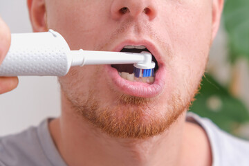 A young man with red beard is brushing his teeth with a white electric brush. Oral care.