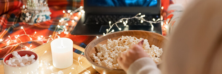 woman's hand popcorn and watching the movie "home alone" on laptop at home in cozy bed with glowing lights garland night on christmas or new year eve. cup with marshmallow. banner