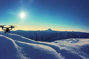 Drone with digital camera flying over winter landscape with snow covered mountains and trees. White remote controlled quadcopter hovering above snowy mountain peak. Close up shot of drone (UAV).
