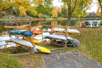 A rack of kayaks on a wooden stand-in a park beside calm water, daytime, fall foliage, nobody