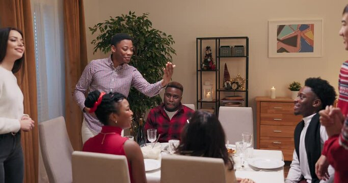 Man of African appearance invited friends to birthday party. Young people are sitting at the table, smiling and talking. His work colleagues entered the room and he introduced friends to each other.