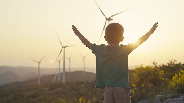 The boy rejoices and waves his hands at the sunset near the windmills. A child and a source of renewable ecological wind energy in the mountains. Production of electricity in a wind farm. 