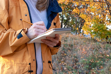Woman in yellow coat holding sketchbook and a pencil on yellow autumn trees background in park....