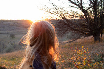A young woman with long blond hair is enjoying the sunset and the autumn landscape. Back view