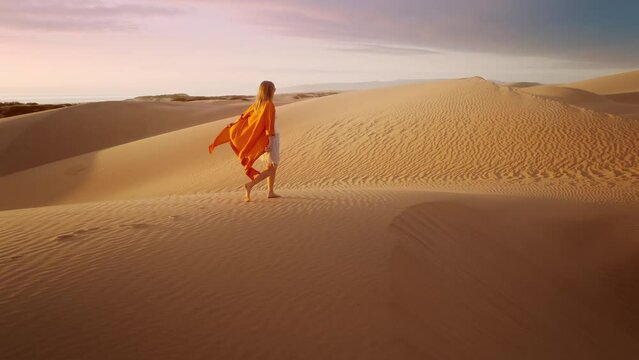 Barefoot female traveler in waving on wind orange dress walking by rippled sand desert surface with dark grey clouds at sunset. 4K Scenic nature aerial. Cinematic slow motion of woman hiking sand dune