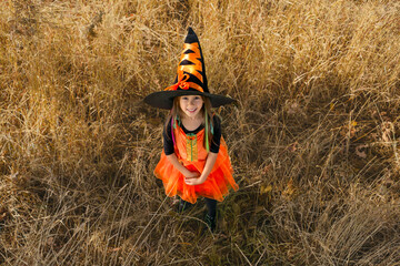 A girl dressed as a witch in a huge black hat with an orange ribbon looks up standing in the tall...