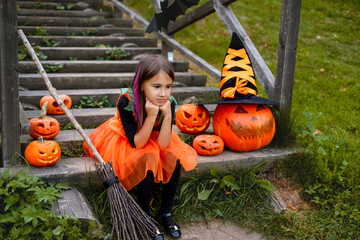 A girl with colored strands in her hair, dressed up as a witch, is sitting on the wooden steps...