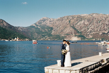 Groom kisses bride on the forehead on the pier by the sea against the backdrop of the mountains