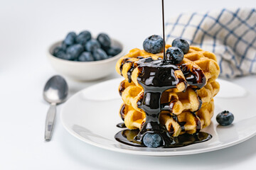 Pouring chocolate syrup on stack of fresh delicious homemade waffles topping with ripe organic blueberry fruit on white ceramic dish .