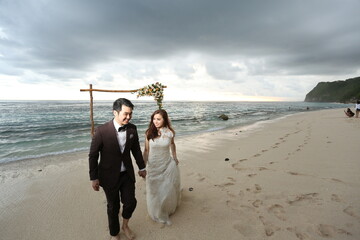 a man in suite and her woman in gown were holding hands, walking together along the beach during...
