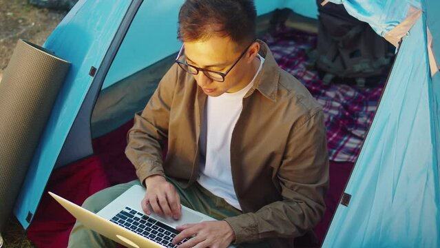 Asian man tourist using laptop computer in campground. Young hiker resting in tent, forest camp, traveling and hiking alone. Summer tourism, wild nature.