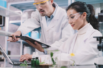 Two research people are comparing information one is working behind a laptop and research man is making notes on a pen and pointing to a laptop in his notebook in a laboratory at a table