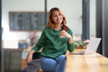 Beautiful young Asian woman relaxing holding coffee and smiling brightly with laptop in cafe.