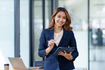 Attractive smiling Asian businesswoman standing holding tablet working and recording work details in office. - Powered by Adobe