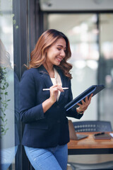 Fototapeta premium Attractive smiling Asian businesswoman standing holding tablet working and recording work details in office.
