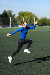Fototapeta na wymiar Women and sport. Smiling Girl in sportswear - blue shirt and black leggings does exercises: bouncing and having fun on the grass at the stadium outdoor on a sunny day. Middle aged sportswoman