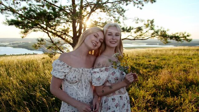 Two cute blonde girlfriends in identical dresses hold flowers and posing for the camera on the field