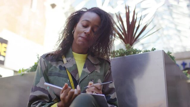 Portrait Of Young Woman Writing In A Notebook As She Sits On A Bench Outside