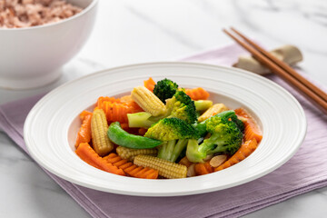 Stir-fried assorted vegetables like broccolis, baby corns, sugar snap peas can carrots, a Thai Chinese dish called pad pak ruam, served on a white plate together with a bowl of brown jasmine rice.