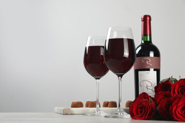 Bottle with glasses of red wine, beautiful roses and chocolate candies on white table. Space for text