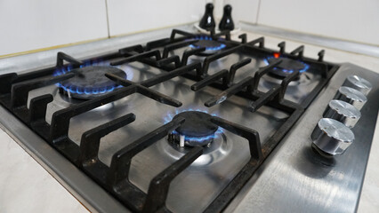 A blue fire is burning on the gas stove. Gas at home. Metal gas stove. Black bars. Light switches of burners. The gas is working. Beige tiles on the walls. Stop the economic crisis. Warmth in house