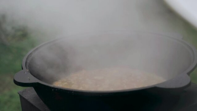 The cook lifts the lid and stirs the gravy for pilaf in the cast-iron cauldron with a slotted spoon. Close-up.