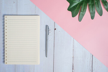 Top view image of notebook with pen and xanadu leaf on wooden table with pink background