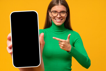 Happy young woman pointing to blank phone screen, isolated on yellow background
