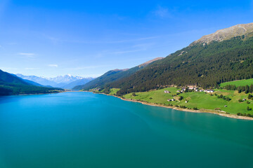 Fototapeta na wymiar Aerial view of lake (Reschensee). Large reservoir surrounded by mountains at sunny noon. Recreation area for tourists and sportsmen. Organic farm on the shore. Italy, Vinschgau, Giern.