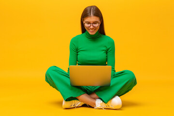 Portrait of woman in green clothes sitting on floor with laptop on knees on yellow background