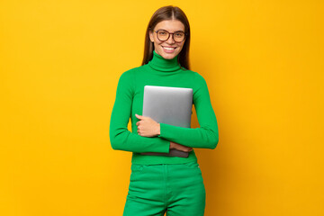 Portrait of smiling female student in green holding laptop, isolated on yellow background