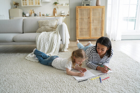 Mom or babysitter and little preschooler kid girl daughter painting together lying on floor carpet