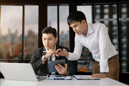 Asian business colleague business presents and explains and using laptop and tablet. Teamwork, financial marketing team,   while sitting in modern office room...