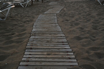 Fototapeta premium Detail view on wooden sidewalk or footpath in sandy beach in a holiday resort during sunrise. In upper left corner are visible plastic deck chairs. In the sand are foot imprints.