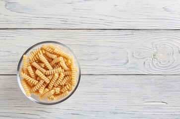 Uncooked fusilli pasta in glass bowl on white wooden background with copyspace
