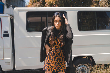 A chic tourist wearing a black leather jacket and a pattern printed short dress with a big gold belt posing outdoors with a white van as background.