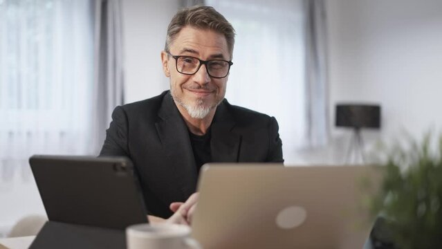 Businessman working with tablet and laptop computer in home office. Happy middle aged, mid adult, mature age man smiling. Entrepreneur sitting at desk, managing business online.