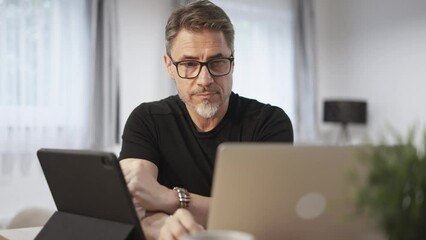 Older businessman working online with laptop and tablet computer at home sitting at desk in living room. Home office, browsing internet. Portrait of happy, mature age, middle age, mid adult man in 50s