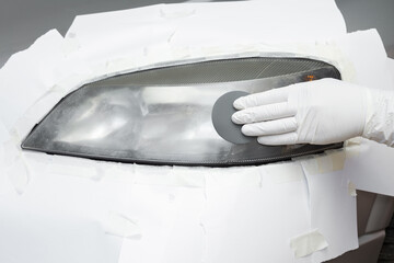 Repairman hand in white rubber protective gloves using sandpaper and sanding car headlight. Renovation process. Restore automobile lamp. Closeup. Front view. © fotoduets