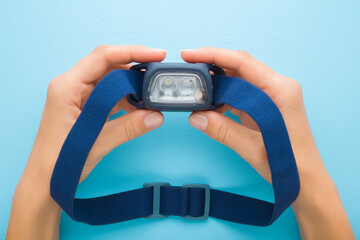 Young adult woman hands holding led headlamp with elastic strap. Light blue table background. Pastel color. Closeup. Equipment for work or sport activities in dark time. Top down view. © fotoduets