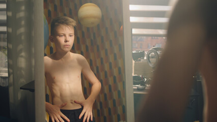 Muscular schoolboy examines his body after weight training exercise in front of mirror in his room. Muscular abs and shoulders. Bare upper body. Weight training, athletes, fitness, physical activity.