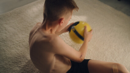 Young enthusiastic boy training abs holds a football while exercising alone indoors. Prepares before the soccer match. Sport and active lifestyle concept.