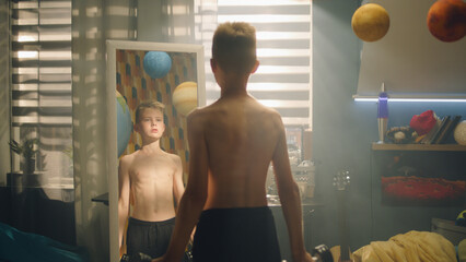 A focused school boy do dumbbells curls stands in front of the mirror in his bedroom. Nude upper body and black shorts. Prepares for a match. Fitness, weight traning concept.
