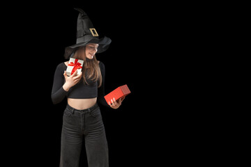 Smiling girl in black clothes and pointed hat holds an open gift. Looks at box in surprise. Witch girl isolated on black background.