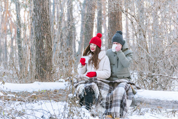 Young happy couple hugging each other in a snow-covered winter park, wrapped in a blanket and drinking tea from mugs.
