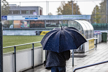 Rear view of a man walking with an umbrella on rainy day next to a soccer field in Dutch village, drops falling, wearing blue jeans and a rain jacket, green grass and tribunes in blurred background