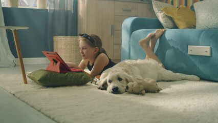 Young girl lying on mild carpet, choosing video on digital tablet computer, dog feeling tired and...