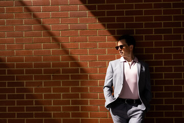 A handsome businessman in a casual grey suit standing half in the shadows and half in the bright sun light against a red brick wall