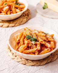 Rustic Plate with penne pasta in red tomato sauce with grated cheese and fresh basil on a table with white textured linen with a ratan plate chargers
