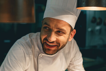 Close up of smiling male chef in uniform standing on kitchen of restaurant and looking away